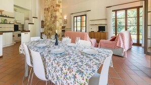 A dining area with a table set for four, a stone wall, a television, and a view of greenery through large windows. - Villa Anna in Bettona, Perugia, with Swimming Pool and Tennis Court (Bettona)