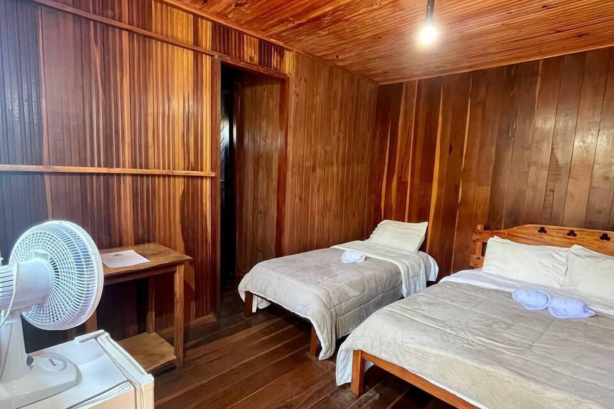 A wooden-paneled room with two beds, a bedside table, and a standing fan.