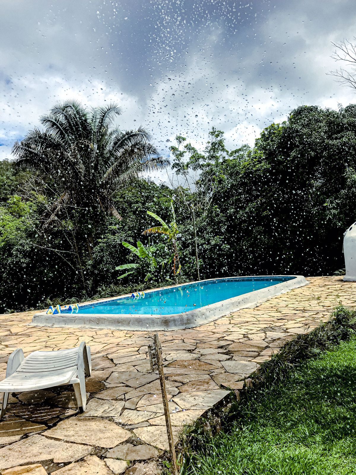Una piscina con solárium de piedra, rodeada de abundante vegetación y un cielo nublado.