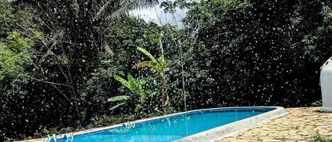 Una piscina con solárium de piedra, rodeada de abundante vegetación y un cielo nublado.