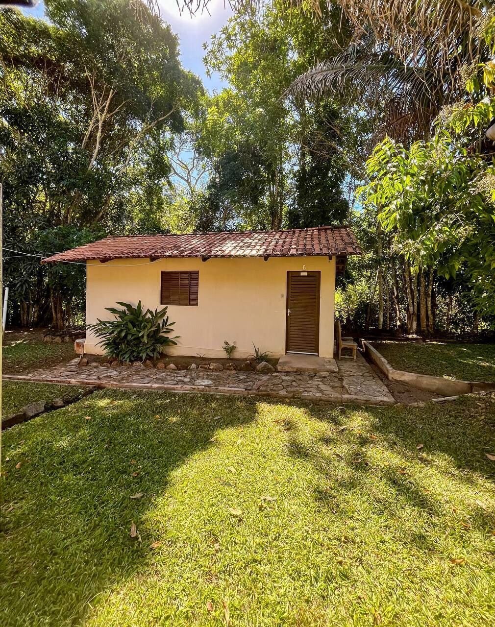 A small, single-story building with a tiled roof, a door, and a small window. It is surrounded by a well-maintained lawn and various plants.