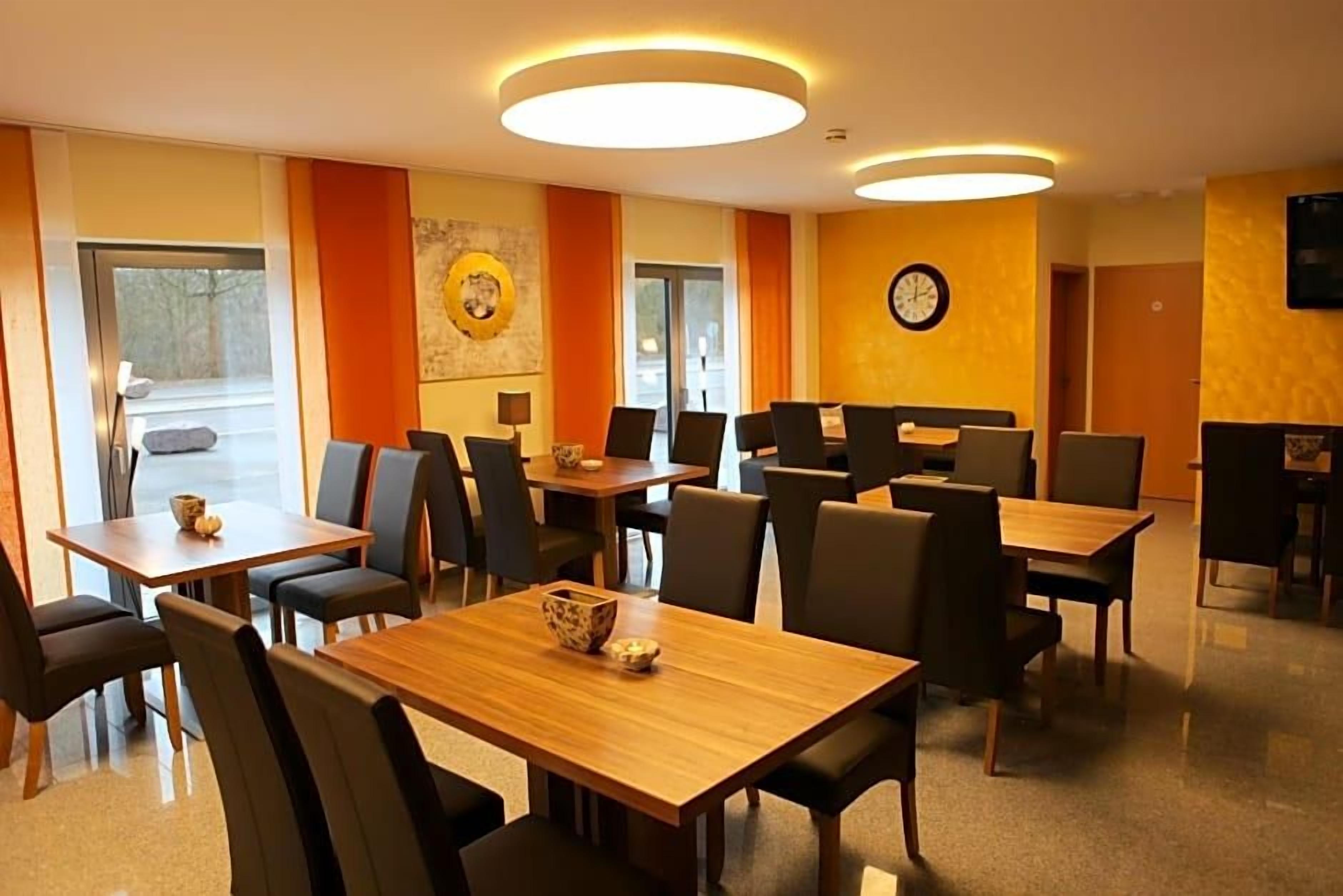 Een restaurant in een hotel met meerdere tafels en stoelen, een klok aan de muur en een raam dat natuurlijk licht laat binnen.