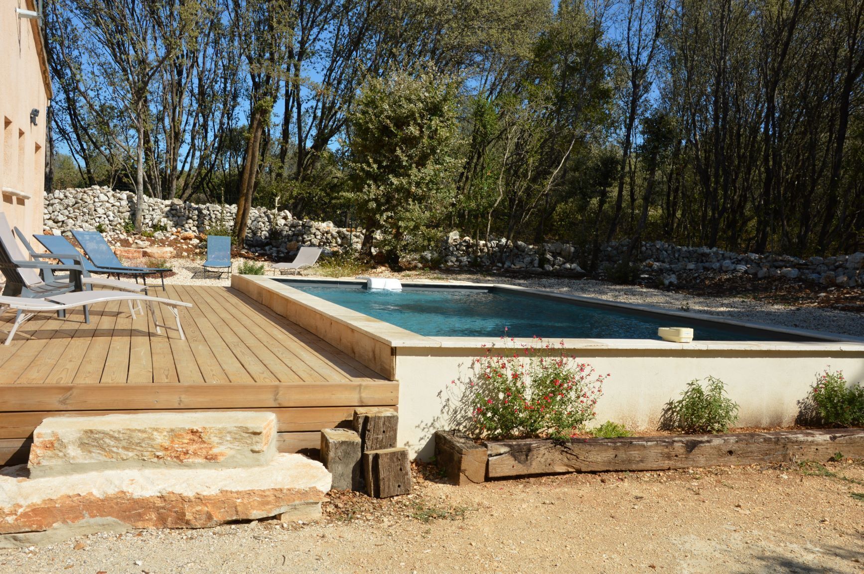 Zona de piscina con muelle de madera, sillones de descanso y pared de piedra.