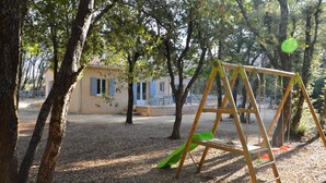 Un terrain de jeux avec des balançoires et un toboggan, entouré d’arbres et bordé d’un bâtiment aux volets bleus.