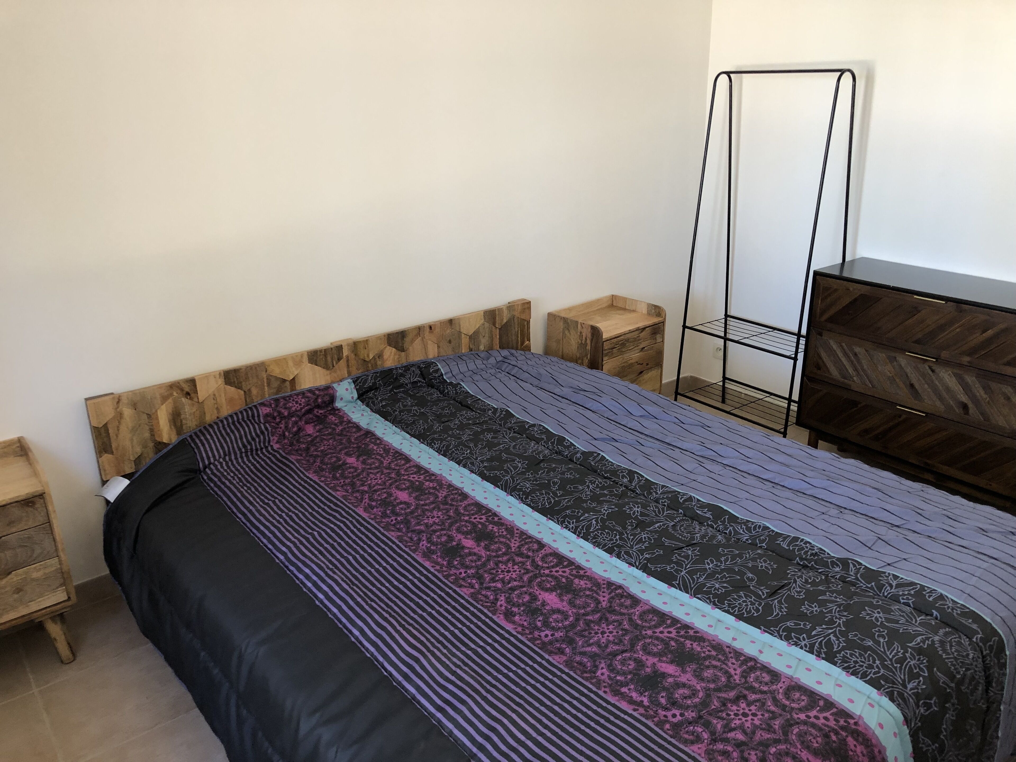 A bedroom with a wooden headboard, a bed with patterned bedding, a nightstand, a dresser, and a metal shelf unit.