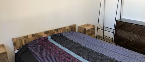 A bedroom with a wooden headboard, a bed with patterned bedding, a nightstand, a dresser, and a metal shelf unit.