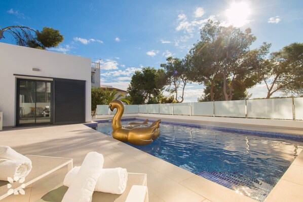 Une piscine avec un cygne flottant, des chaises longues et un bâtiment moderne doté d’une porte en verre.
