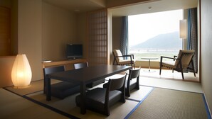 A traditional Japanese room with tatami flooring, a low table with chairs, a television, and a view of the sea.