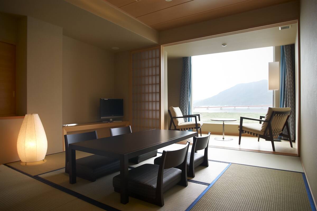 A traditional Japanese room with tatami flooring, a low table with chairs, a television, and a view of the sea.