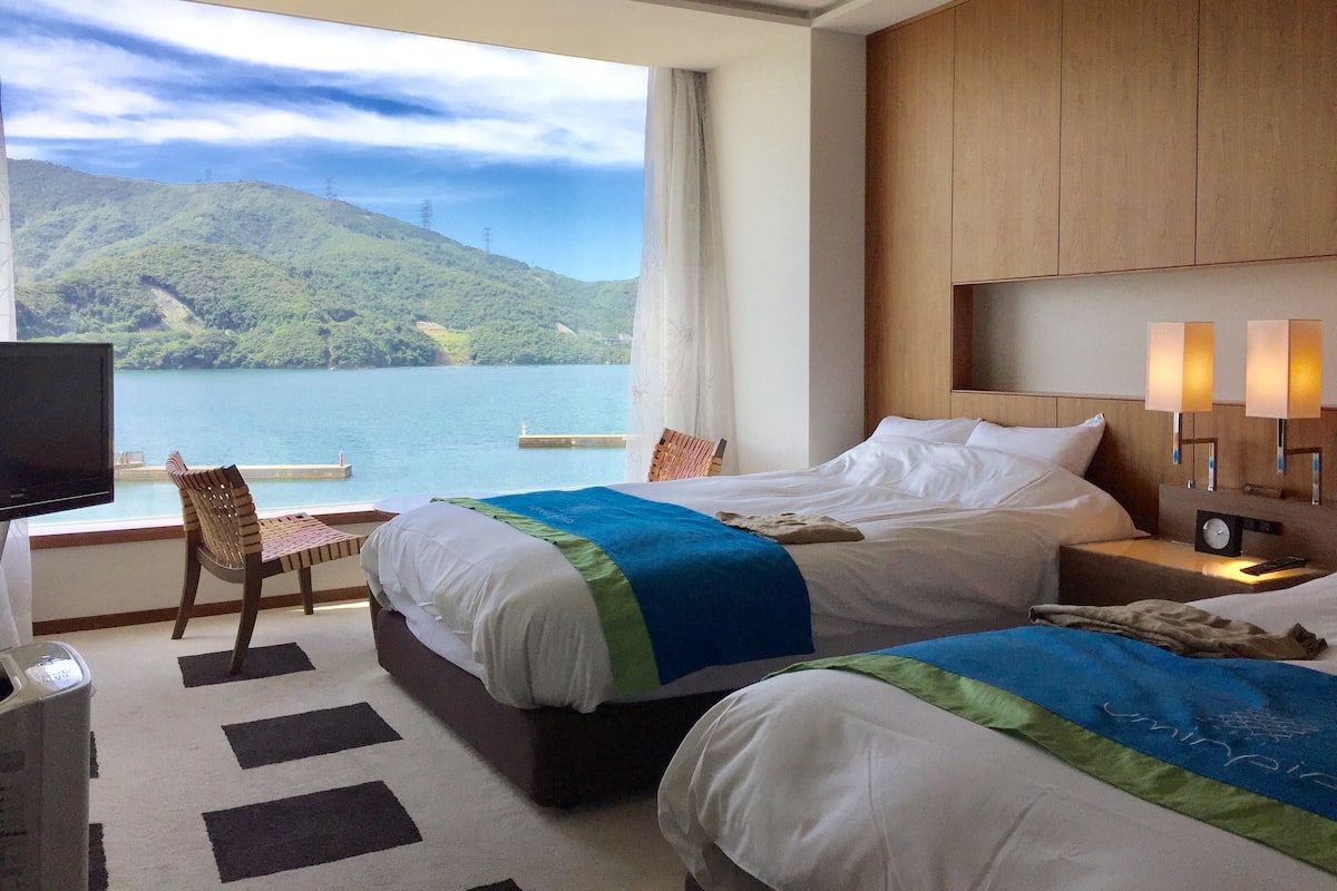 A hotel room with two beds, a TV, a desk, and a view of a lake and mountains.