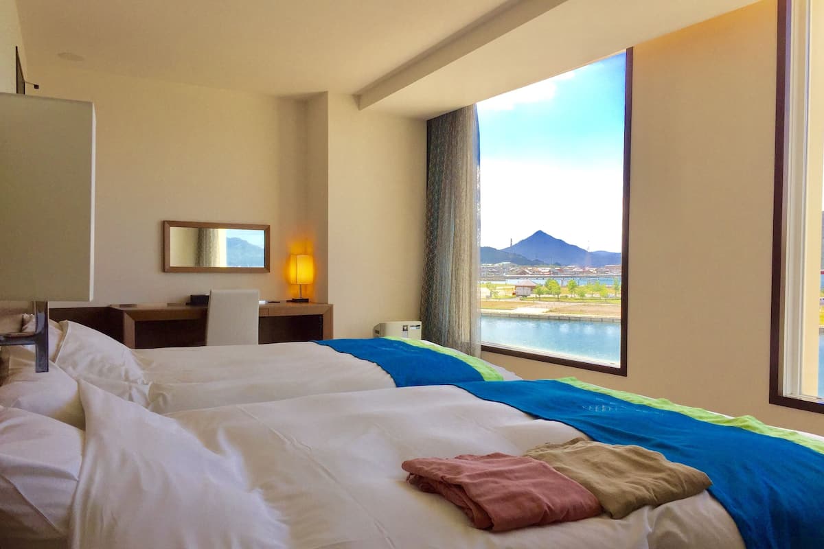 A hotel room with two beds, a desk, a lamp, and a large window with a view of a body of water and a mountain.