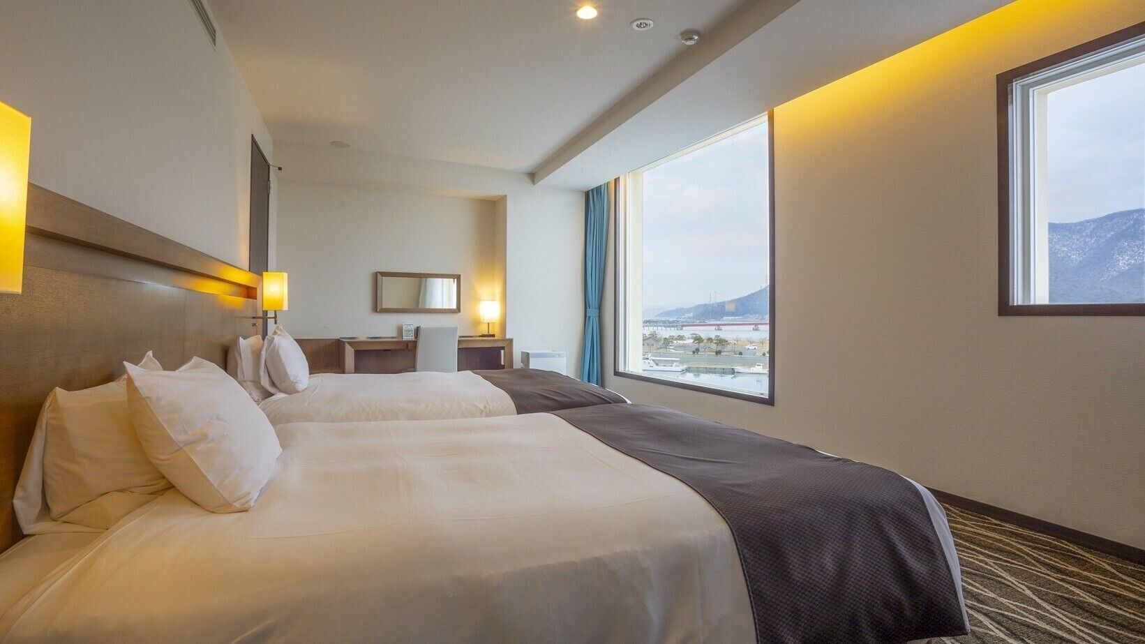 Habitación de hotel con dos camas, un escritorio, una silla, un ventanal con vistas y un espejo en la pared.
