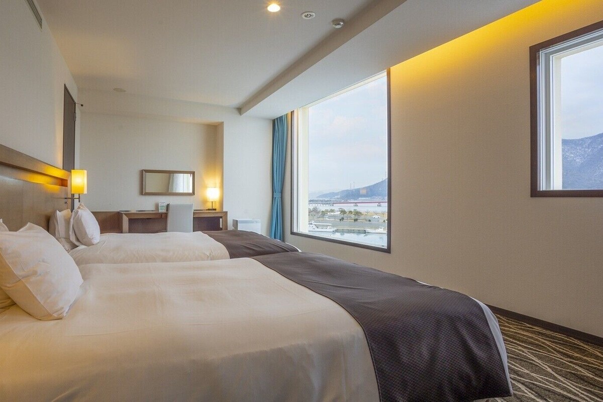 A hotel room with two beds, a desk, a chair, a large window with a view, and a mirror on the wall.