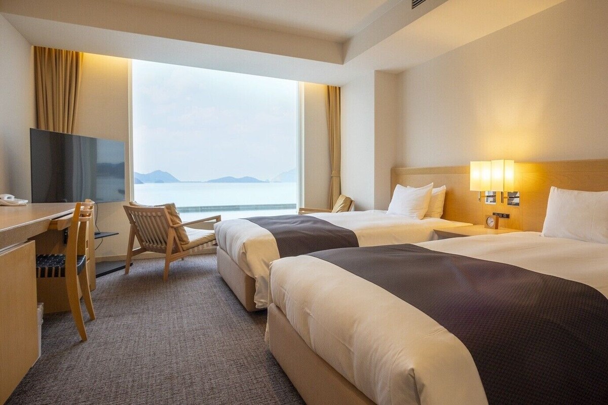 Habitación de hotel con dos camas, un escritorio con silla, un televisor y vista al mar.