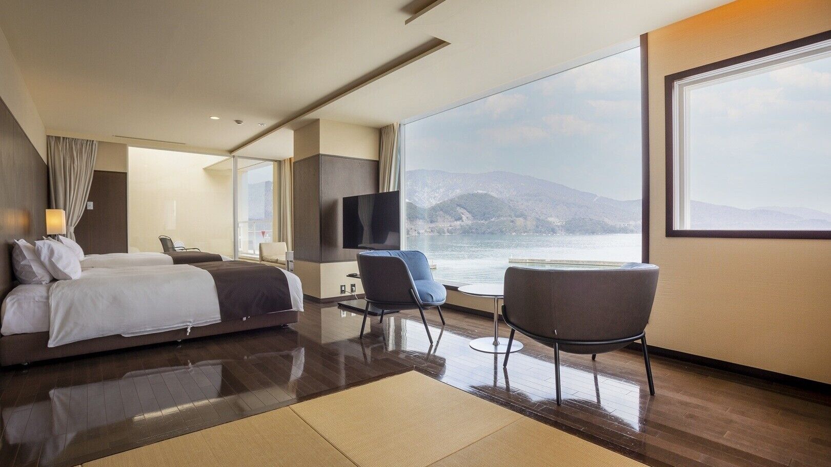 Una habitación de hotel moderna con una cama grande, un televisor, un área de descanso con vista al mar y un ventanal amplio.