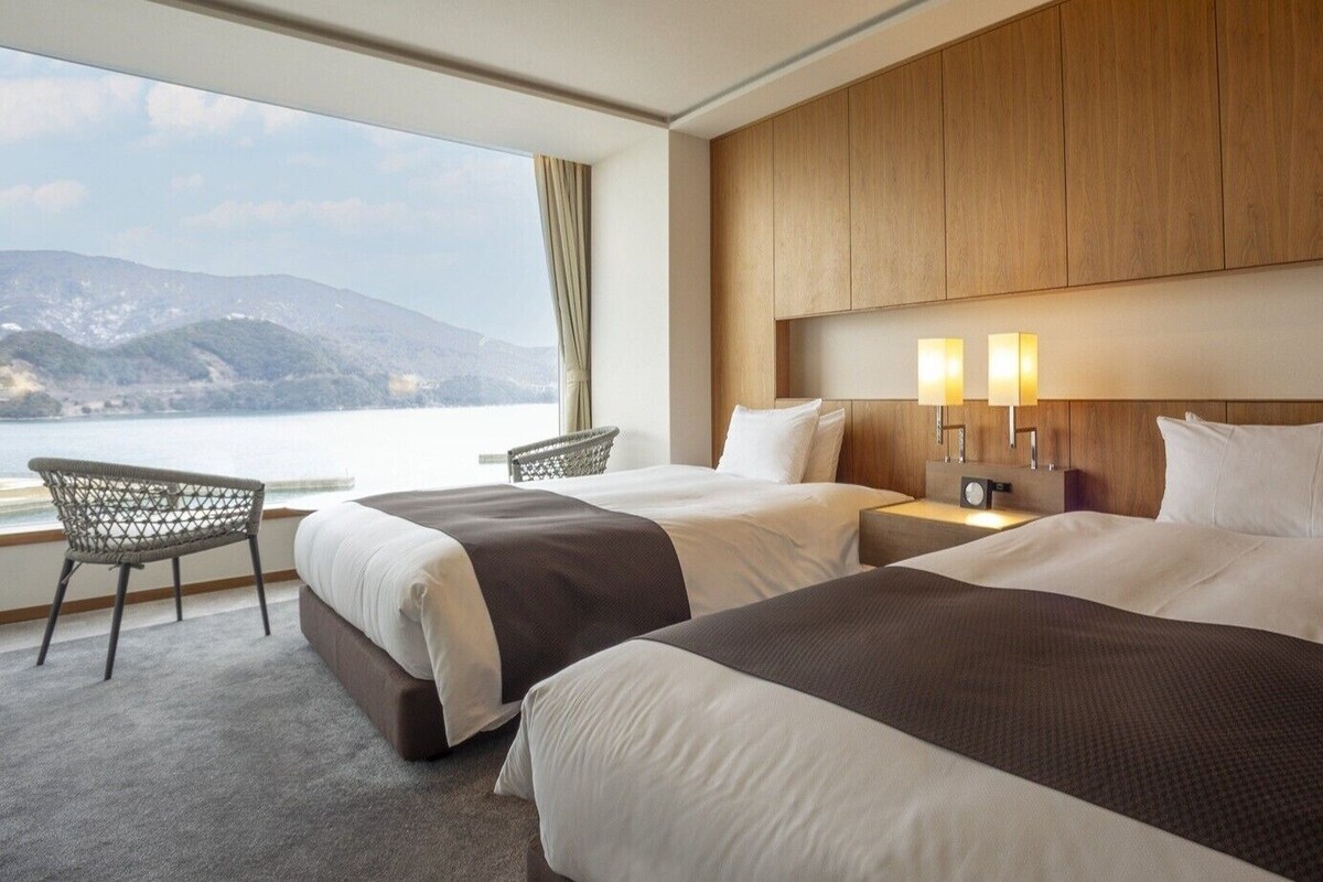 Habitación de hotel con dos camas, vista al mar, cabecero de madera y una silla junto a la ventana.