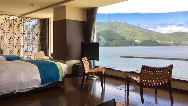 A hotel room with a bed, two wicker chairs, a small table, a TV, and a large window overlooking a lake and hills.