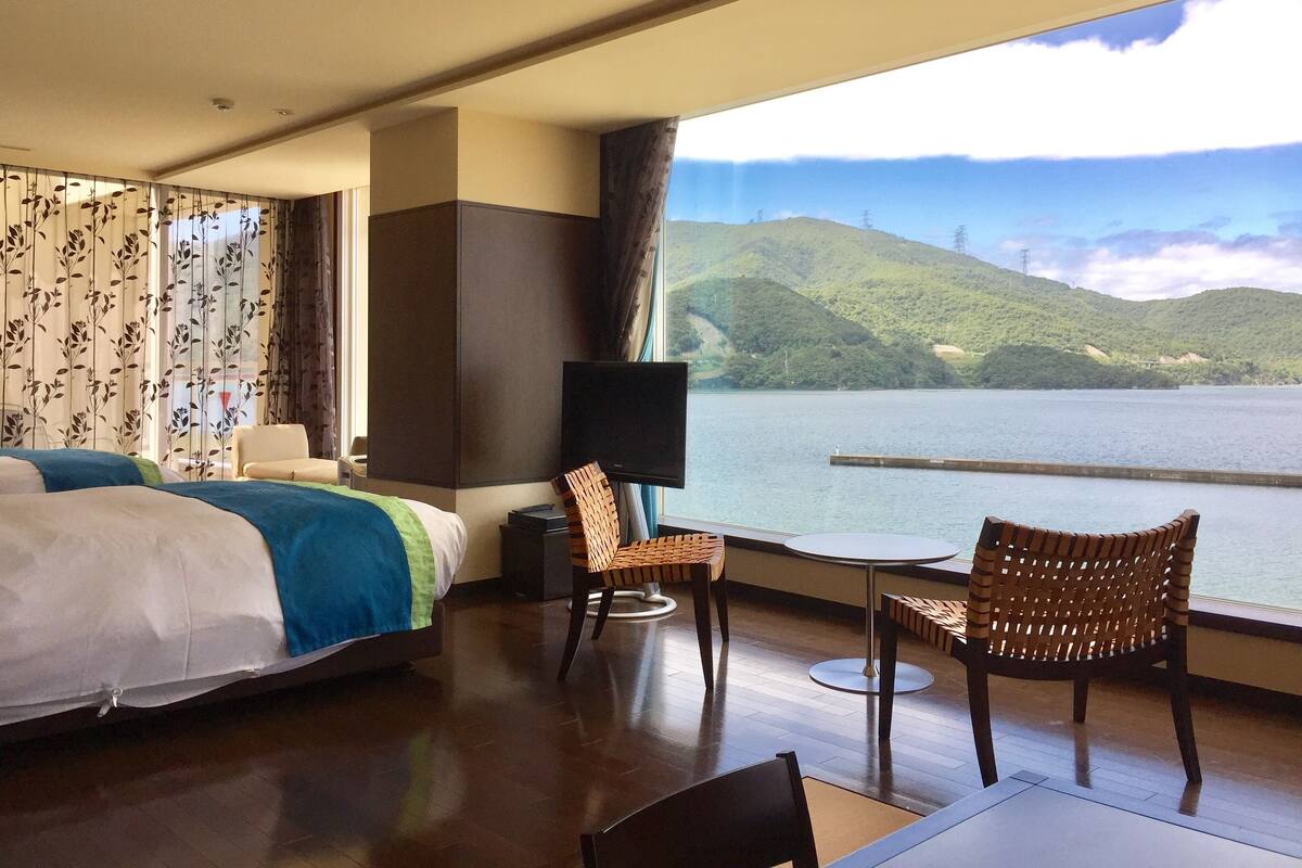 A hotel room with a bed, two wicker chairs, a small table, a TV, and a large window overlooking a lake and hills.