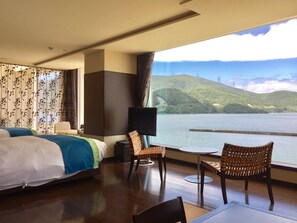 A hotel room with a bed, two wicker chairs, a small table, a TV, and a large window overlooking a lake and hills. - Ocean Resort Hotel & Spa Uminpia (Ooi)