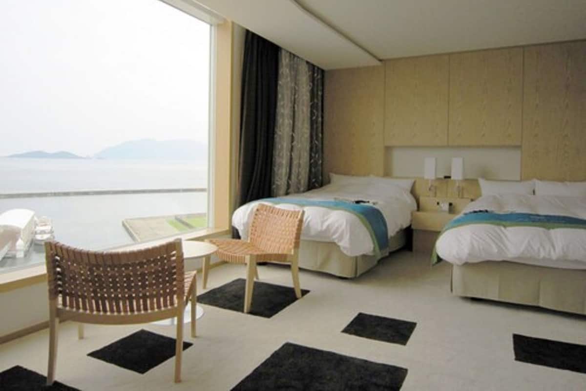 A hotel room with a large window, two beds, a desk with a chair, and a view of the sea.