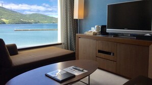 A hotel room with a large window offering a view of a lake and hills. There's a TV on a wooden cabinet, a sofa, and a small table with a remote control and a book.