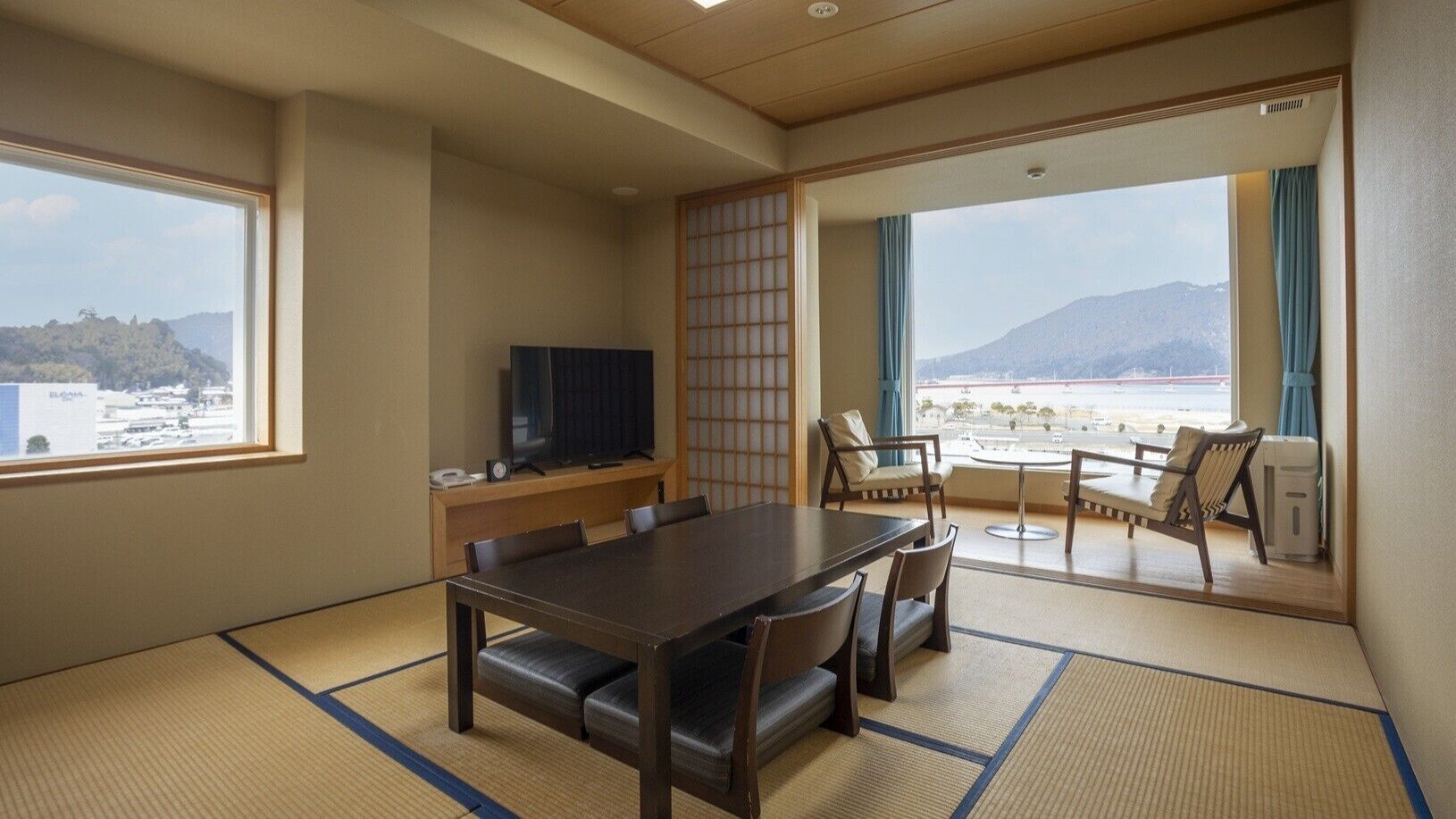 Una habitación con suelo de tatami, una mesa de comedor con sillas, un televisor y vistas a un paisaje montañoso a través de ventanas grandes.