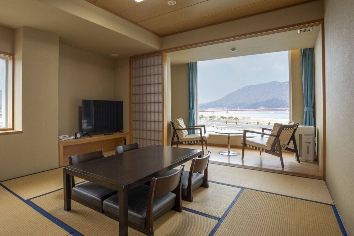 A room with tatami flooring, a dining table with chairs, a television, and a view of a mountainous landscape through large windows.