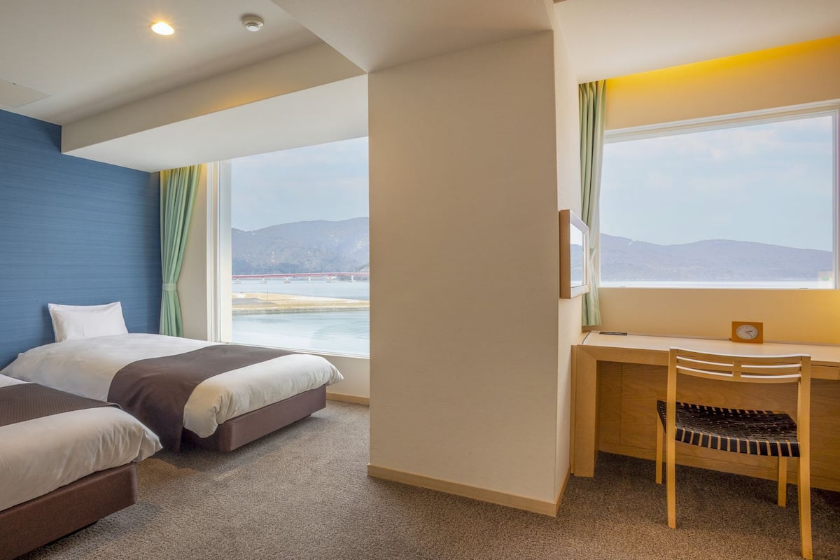 A hotel room with two beds, a desk, and a chair. There is a large window with a view of a body of water and mountains.