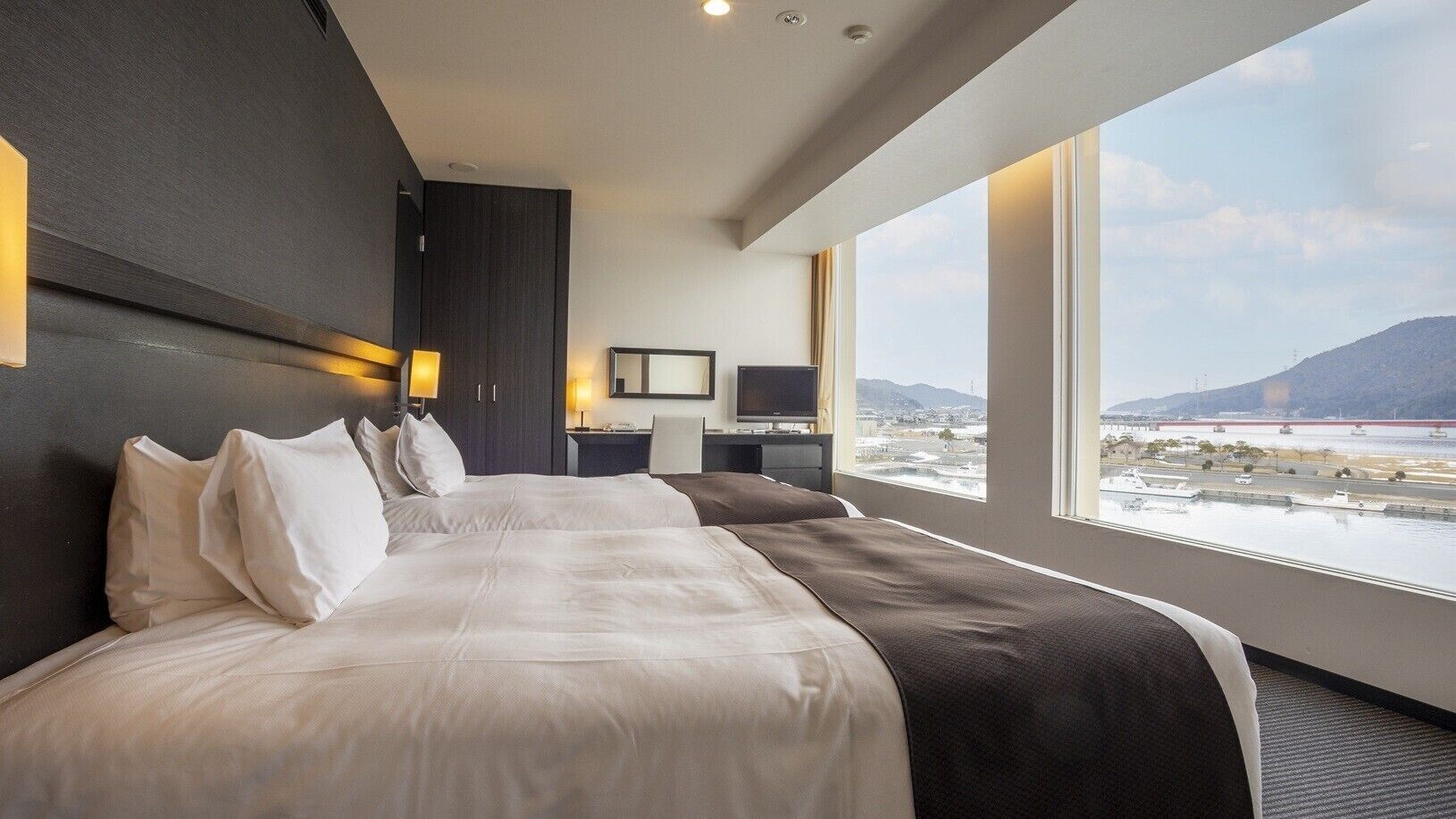 Habitación de hotel con dos camas, un escritorio, una silla, un televisor y un amplio ventanal con vistas al agua y montañas.