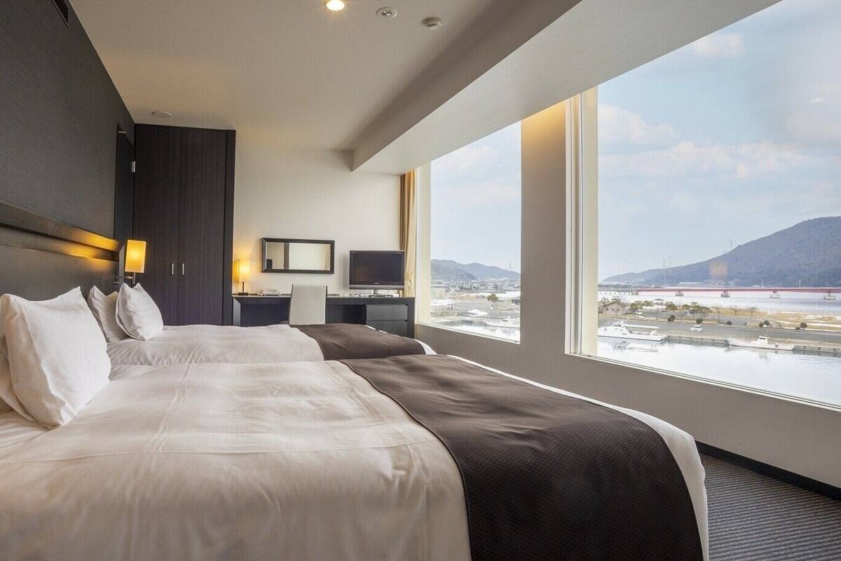 A hotel room with two beds, a desk, a chair, a TV, and a large window with a view of a body of water and mountains.