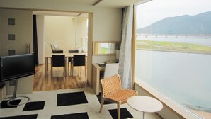 A modern hotel room with a large window offering a view of a bridge and water. There is a dining area with a table and chairs, and a small seating area with a chair and a table.