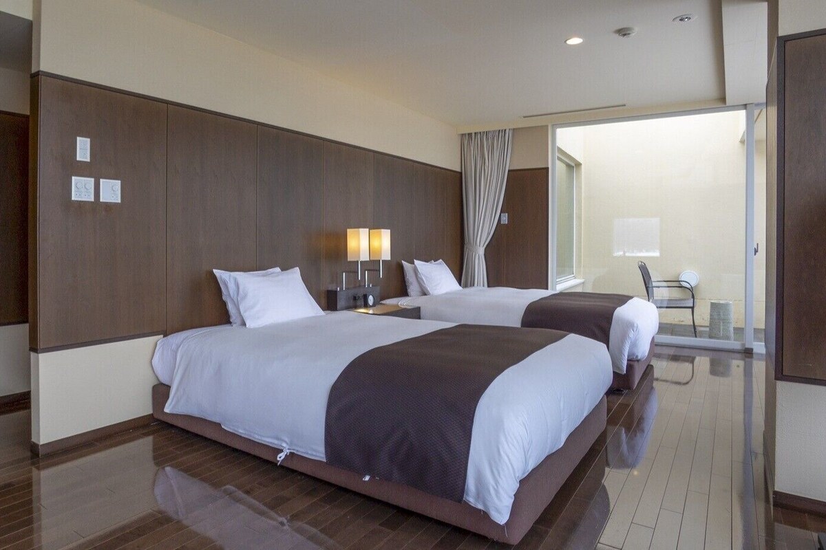 Habitación de hotel con dos camas, una mesita de noche con lámpara, una silla y un ventanal corredero a un balcón.