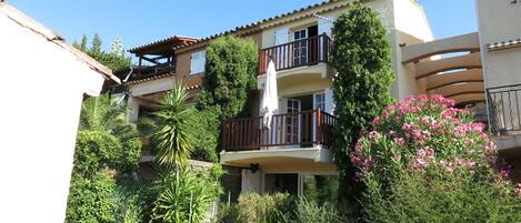 A multi-story residential building with balconies, surrounded by lush greenery and flowering plants.