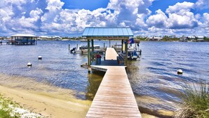 A wooden pier leading to a covered pavilion with a blue roof, surrounded by calm water and distant houses. - Sundown B 13 (Pensacola)