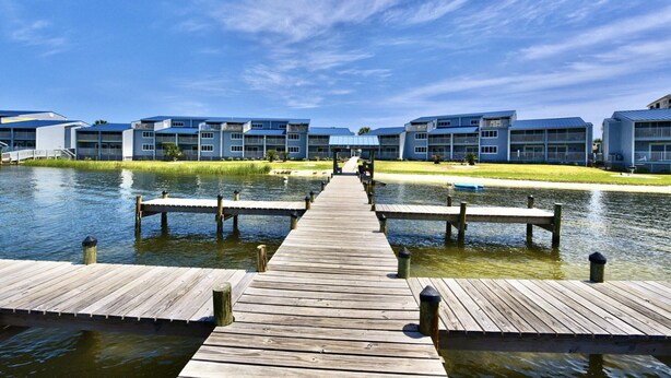 A wooden pier leading to a row of modern buildings with balconies, over a calm body of water. - Sundown B 13 (Pensacola)