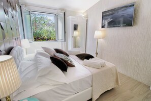A hotel room with a bed, bedside lamps, a window with a view of greenery, a mirror, and a framed picture on the wall. - Villa Magdala - Three Bedroom Villa, Sleeps 8 (La Maddalena)