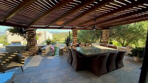 An outdoor patio with a pergola, stone columns, wicker furniture, and a glass-top table. - Villa Magdala - Three Bedroom Villa, Sleeps 8 (La Maddalena)