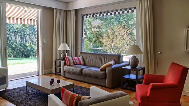 Una sala de estar moderna con un sofá, sillones y una mesita de centro. Hay dos lámparas sobre mesitas auxiliares y un ventanal con cortinas.