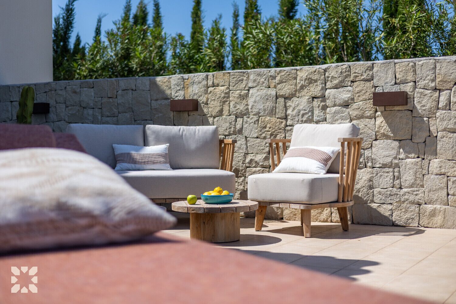 Un patio con pared de piedra, un sofá, dos sillones y una mesa redonda de madera con un cuenco de fruta.