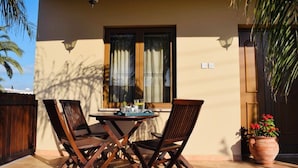Une terrasse avec une table et quatre chaises, une fenêtre avec des rideaux, une plante en pot et une porte avec une plante placée à côté.