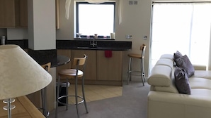 A modern kitchen with a central island, bar stools, a dining table, and a sofa with cushions. - Beachside, stunning apartment with sea and harbour views (West Bay, Bridport)