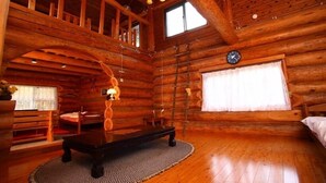 Interior de una cabaña de troncos con piso de madera, una mesa de comedor, un banco, una lámpara colgante, un reloj y una ventana con cortinas.