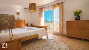 Ein Schlafzimmer mit Bett, Holzkommode, Korbsessel und Blick ins Freie.