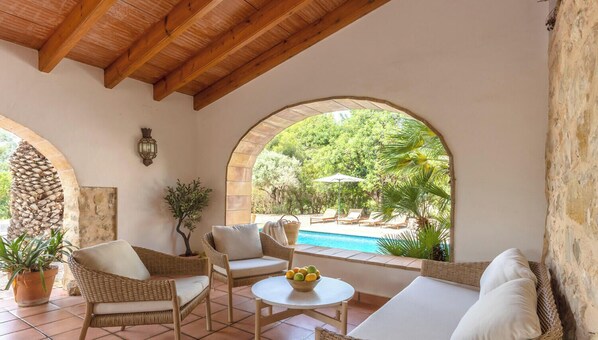 Un espacio de estar techado al aire libre con mobiliario de mimbre, una pequeña mesa con fruta y vistas a una piscina y follaje.