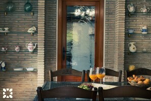 A dining area with a brick wall, glass shelves with tea sets, and a table with chairs and fruit.
