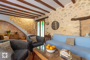 A cozy living room with stone walls, a blue sofa, two armchairs, a glass coffee table with a bowl of lemons, and a staircase with a stone arch.