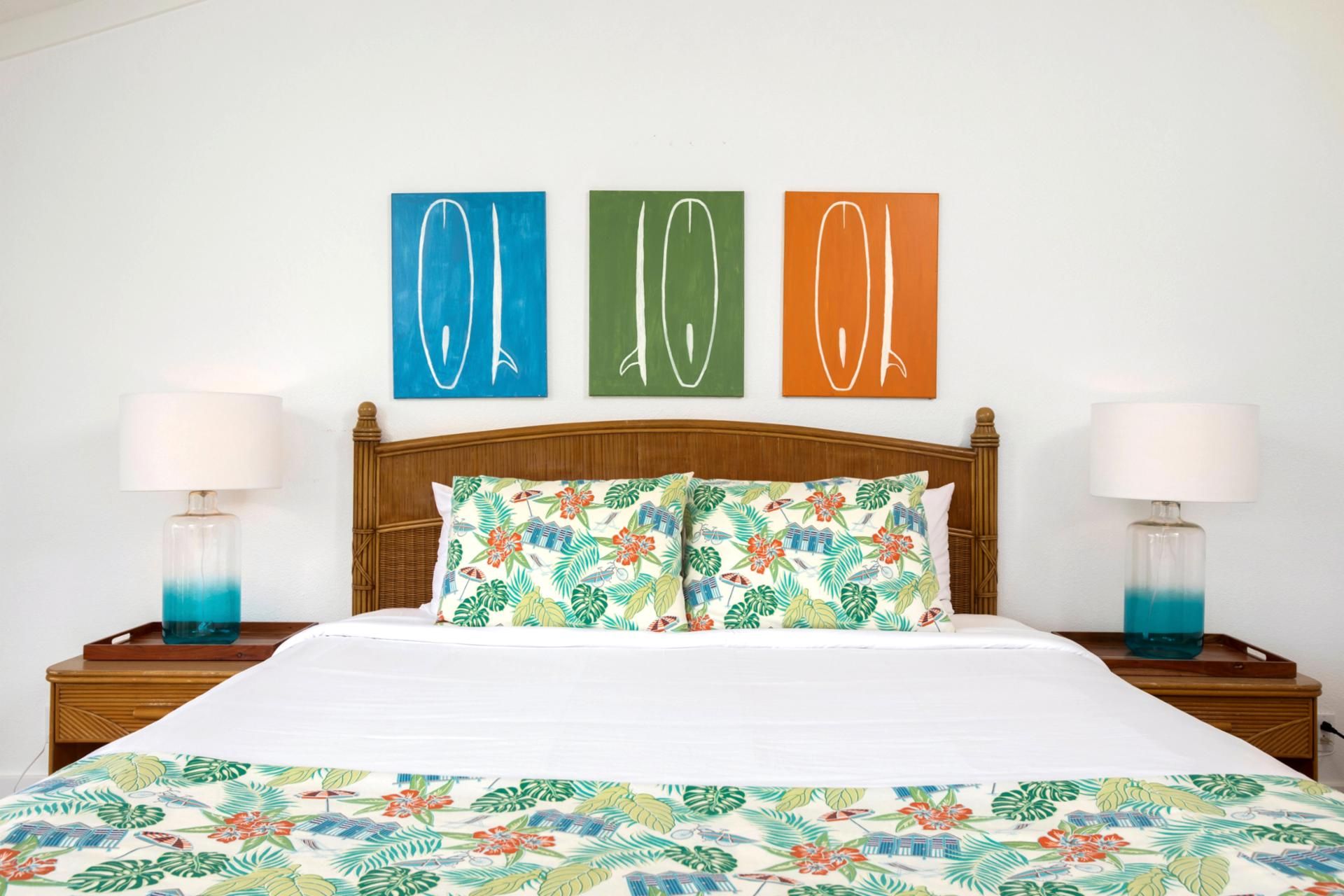 Ett hotellrum med en sänggavel i trä, ett sängöverkast med blommönster och tre konstverk i blått, grönt och orange som hänger på väggen.