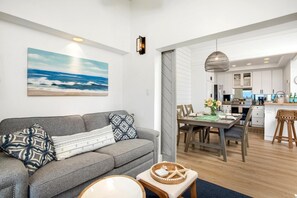 A modern living room with a grey sofa, a dining area with a wooden table, and a kitchen in the background. - Kapalua Golf Villas | Golf View 2 BR | Car Incl. w/ 6+ Nights | KGV-19P2 by KBM (Lahaina)