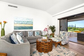 Ein modernes Wohnzimmer mit einem grauen Ecksofa, einem runden Couchtisch und Blick auf einen Golfplatz durch eine Glasschiebetür.