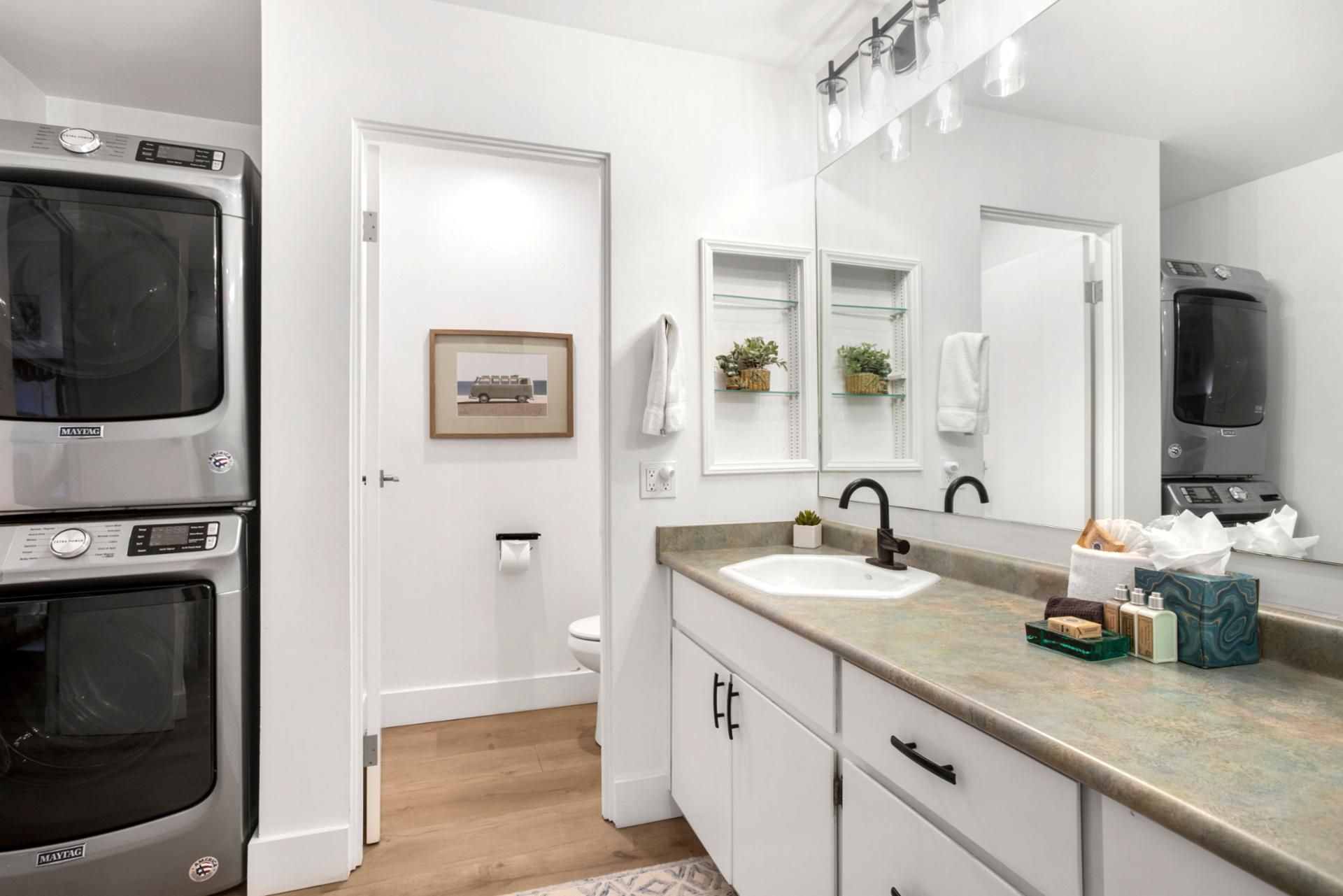 A modern bathroom with a large mirror, a sink, and a toilet. There is a Maytag washer and dryer in the adjacent room.